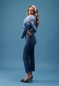 Jeanjacka, blå jeans och svarta högklackade skor. Vågigt hår med ett vitt pannband. Blå bakgrund fulländar looken.