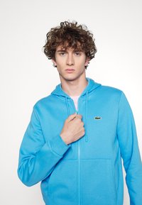 Sudadera azul con cremallera hecha de un tejido suave, que cuenta con un bolsillo canguro y un pequeño logo bordado en el pecho. Incluye capucha con cordones.
