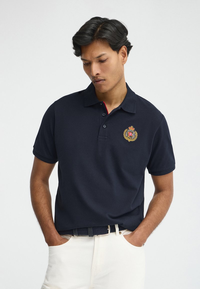 Uomo con polo blu navy con stemma ricamato dorato e rosso sul petto, pantaloni bianchi e cintura nera, mani in tasca.