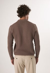 Bruine gebreide trui met een kraag, met lange mouwen. Gepaard met lichtbeige broek, zichtbaar detail van de achterzakken.