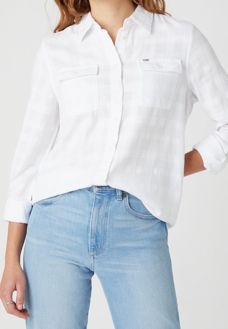 Femme portant une chemise blanche à manches courtes avec poches poitrine et un jean taille haute bleu clair, les manches de la chemise retroussées.