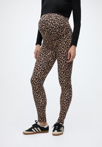 Έγκυος άτομο που φοράει κολάν εγκυμοσύνης με λεοπάρ μοτίβο, μαύρο μακρυμάνικο τοπ και μαύρα-άσπρα αθλητικά παπούτσια, στέκεται σε λευκό φόντο.