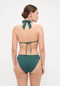 Tumši zaļš bikini komplekts ar kakla saiti un loku, kā arī augsto vidukli, izgatavots no gludas, stiepļu auduma.