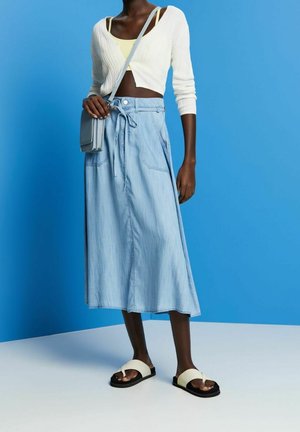 Světle modrá džínová sukně s uvázaným pasem a dvěma předními kapsami, doplněná bílým žebrovaným zkráceným kardiganem a krémovými sandály.