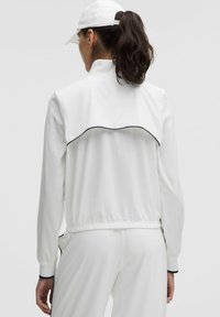 Valge sportlik jakk kõrge kaelusega ja kogumisega vöökoha juures. Omab tagaosa flappi ja kontrastset musta kitsa äärt õlgade juures.