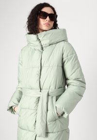 Lys grøn pufferjakke med høj krave, bælte i taljen og quiltet tekstur. Har skjulte lommer og bæres med oversized solbriller.