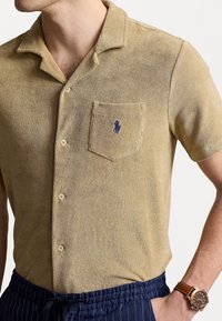 Man i beige kortärmad t-shirt i frotté med krage och knäppning fram, marinblå randiga byxor och brun läderarmband.