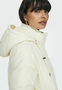 Chaqueta tipo plumón en suave tela crema con capucha, cordón ajustable y textura lisa. Presenta un cierre de cremallera y discretos detalles de costura.