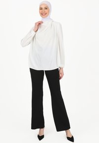 Hvid langærmet bluse, afslappet pasform, draperet halsudskæring, parret med sorte brede bukser og spidse sorte hæle. Model med en hvid hijab.