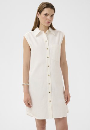 Kobieta ubrana w białą, bezrękawną sukienkę zapinaną na guziki z kołnierzykiem i przednią kieszenią, stojąca na jednolitym tle.