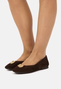 Piernas que llevan zapatos planos de ante marrón oscuro con puntas afiladas, cada uno adornado con un gran acento decorativo irregular dorado en la parte superior.