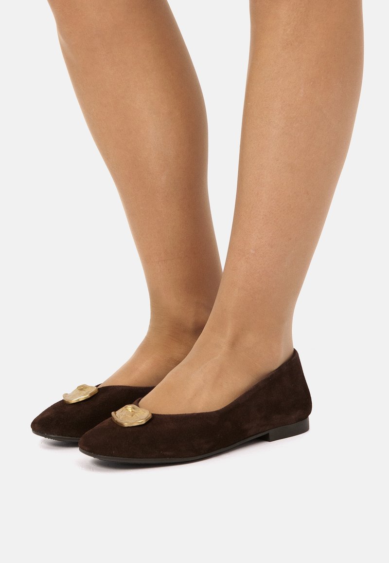 Piernas que llevan zapatos planos de ante marrón oscuro con puntas afiladas, cada uno adornado con un gran acento decorativo irregular dorado en la parte superior.