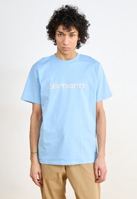 Light blue cotton t-shirt featuring a prominent beige logo print on the front, short sleeves, and a crew neckline. Smooth texture.