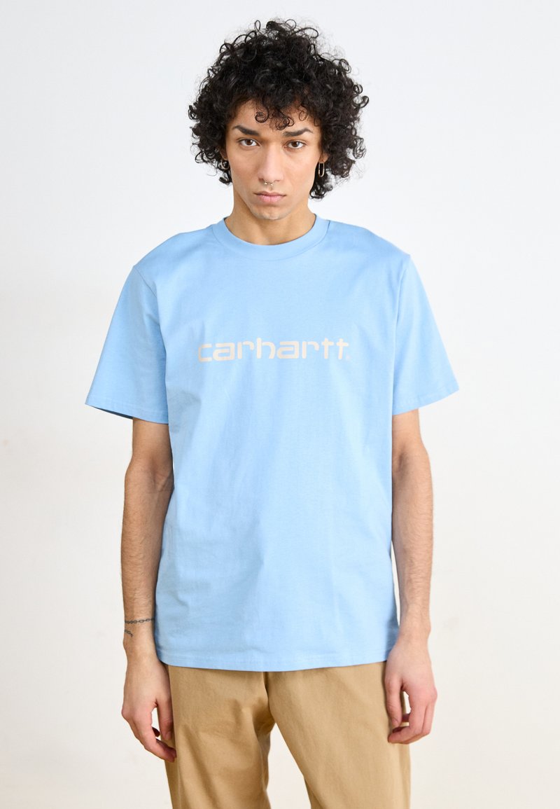 Light blue cotton t-shirt featuring a prominent beige logo print on the front, short sleeves, and a crew neckline. Smooth texture.