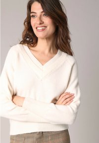 Femme aux cheveux bruns ondulés portant un pull blanc en V et un pantalon à carreaux marron, souriante et les bras croisés devant un fond uni.