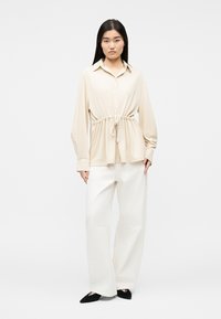 Žena s dlhými tmavými vlasmi, oblečená v béžovej blúzke s dlhými rukávmi a zapínaním na gombíky, so zaväzovaním v páse, bielych širokých nohaviciach a čiernych špicatých topánkach stojaca.