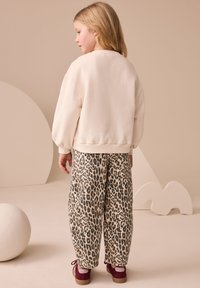 Bluză sport de culoare bej deschis cu mâneci bufante, combinată cu pantaloni largi cu imprimeu de leopard crem și negru, completată de pantofi vișinii.