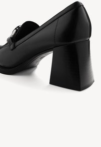 Černá kožená botička na vysokém podpatku s masivním blokovým podpatkem a dekorativním páskem vpředu. Hladká textura a štíhlý design.