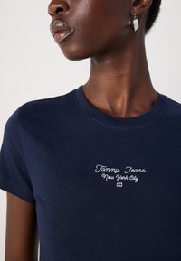 Marineblå bomull T-skjorte med korte ermer, med hvit brodert tekst "Tommy Jeans New York City" på brystet. Rund hals.