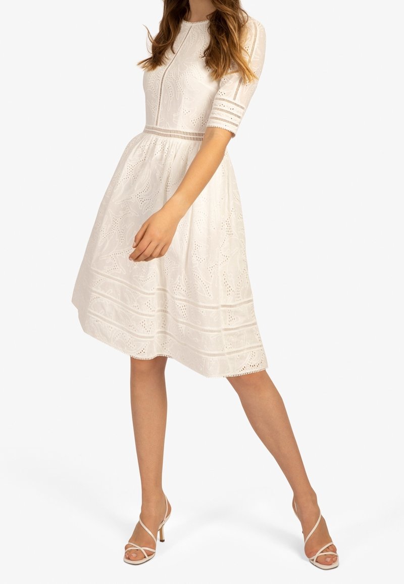 Vestido blanco de manga corta con patrones de eyelet florales, un cuerpo ajustado y una falda acampanada. Combinado con sandalias de tiras beige.