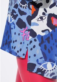 T-shirt med korte ærmer i et blåt stof med abstrakte mønstre, herunder øjne og pletter; pink akcent ved sidesplitten.