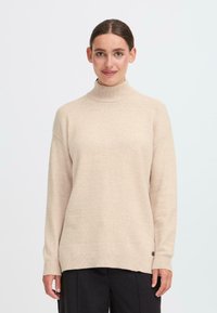 Sieviete, tērpusies beidzīgā turtleneck džemperī un melnās biksēs, stāv uz vienkrāsaina balta fona, vērsta uz priekšu ar nelielu smaidu.