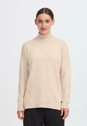 Femme portant un pull à col roulé beige et un pantalon noir, se tenant devant un fond blanc uni, face à l'objectif avec un léger sourire.