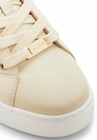 Sneaker in pelle beige con punta arrotondata, superficie testurizzata, lacci piatti e dettaglio metallico dorato con il nome del marchio. Suola bianca.