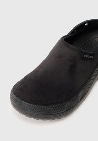 Crne slip-on Crocs cipele s gornjim materijalom nalik na antilop, teksturiranom potplatom i ventilacijskim otvorima na remenu za petu, kosi pogled na bijeloj pozadini.