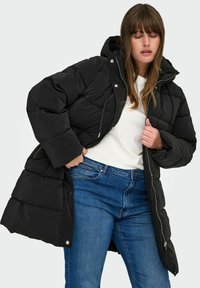 Melnā pūkainā jaka ar jaku; ar quilted dizainu, snap pogām un priekšējo rāvējslēdzēju. Apvienota ar ziliem džinsiem un baltu T-kreklu.
