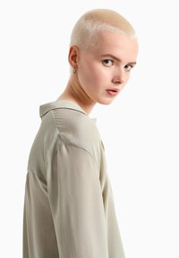 Blusa verde chiaro con colletto, dotata di maniche trasparenti e una consistenza liscia. Il modello ha i capelli corti di un biondo chiaro ed è leggermente girato.