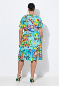 Kleid mit kurzen Ärmeln aus blauer Stoff mit lebhaften gedruckten Mustern, darunter Obst, Gebäude und Blumen. Ausgestellter Saum mit Falten. Grüne Absätze.