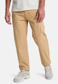 Beige katoenen broek met een rechte pasvorm, voorzien van zijzakken en een soepele textuur, gecombineerd met lichtgekleurde sneakers.
