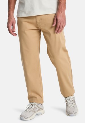 Beige cotton pants with straight fit, featuring side pockets and a smooth texture, paired with light-colored sneakers.
