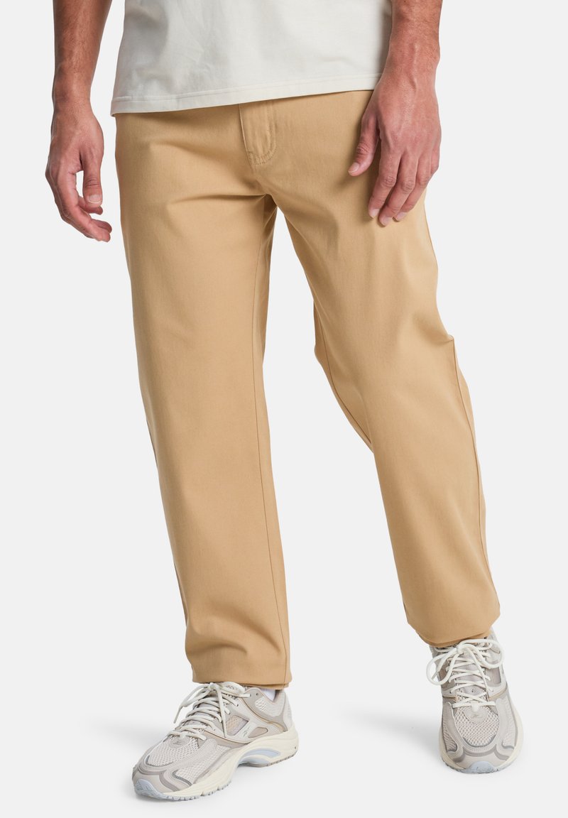 Beige katoenen broek met een rechte pasvorm, voorzien van zijzakken en een soepele textuur, gecombineerd met lichtgekleurde sneakers.