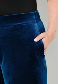 Mörkblå sammetbyxor med mjuk textur, med en bred midje och sidofickor, sedd från midjan till fickan.