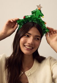Green sequin Christmas tree headband with tinsel, small colorful pom-poms, and a gold star on top. Worn by a woman with long brown hair.