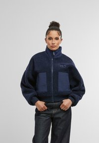 Marinblå fleecejacka med hög krage, dragkedja och stora lappar. Har en texturerad yta och ribbad nederkant. Matchas med mörka jeans.