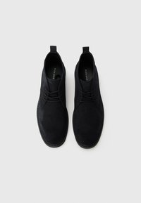 Zapatos de ante negros con cordones y punta redondeada, con tiradores y suela plana. Diseño limpio, textura suave y acentos minimalistas.