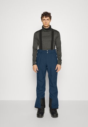 Marineblaue Skihosen mit schwarzen Akzenten, verstellbaren Trägern, Frontknöpfen und geradem Schnitt. Getragen mit einem grauen Langarmoberteil.