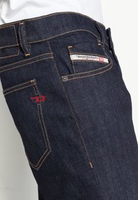 Jeans de mezclilla azul oscuro con costuras naranjas, que presentan una etiqueta de marca en el bolsillo trasero y un acento de logo rojo en el bolsillo lateral.