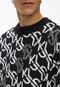 Melna adīta džemperis, kuram ir atkārtots balts logo raksts, ar ribotu apkakli un mīkstu tekstūru. Dizainā ir iekļauti leņķi, ģeometriskas formas.