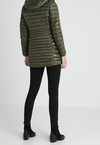 Olivengrøn, polstret jakke med hætte, der har vandrette quiltede linjer og en lille logo detalje ved kanten. Iført med sorte bukser og støvler.