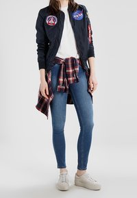 Granatowa kurtka bombowiec z naszywkami NASA, biała koszulka, niebieskie skinny jeans, czerwono-czarna koszula w kratę związana w talii oraz białe sneakersy.