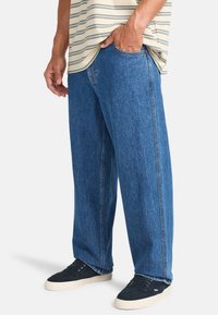 Helle blaue Denim-Jeans mit lockerer Passform, ausgestattet mit einem geraden Bein-Design, dezenten Waschungen und seitlichen Taschen. Getragen mit dunklen Sneakers.