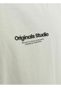 Šviesiai pilkas medvilninis marškinėliai, turintys ryškų juodą tekstą „Originals Studio“. Minkšta tekstūra su paprastu dizainu ir be matomų raštų.