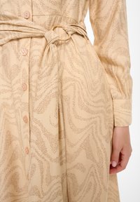 Robe beige avec un motif ondulé, dotée de boutons sur le devant, d'une ceinture à nouer avec un anneau doré et de manches longues. Tissu doux et texturé.