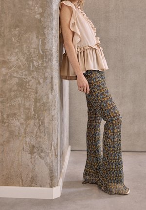 Romige, gerimpelde blouse gecombineerd met aansluitende flared broeken in blauw, bruin en geel met een patroon, tegen een gestructureerde betonnen muur.