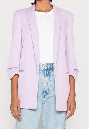 Licht paarse blazer met een op maat gemaakte snit, sjawl kraag, omslagmouwen en geen zichtbare knopen, gecombineerd met een wit shirt en lichte spijkerbroek.