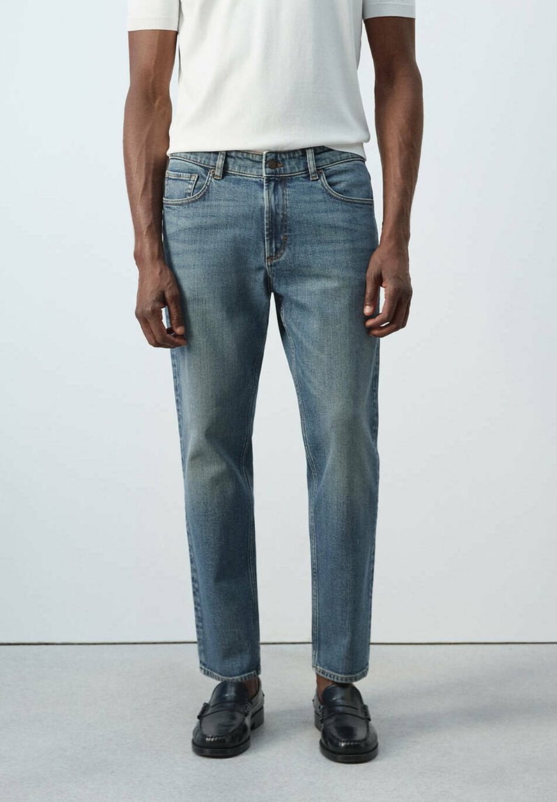 Homme portant un jean bleu délavé, une chemise blanche à manches courtes et des mocassins en cuir noir, debout sur un sol gris contre un mur blanc.